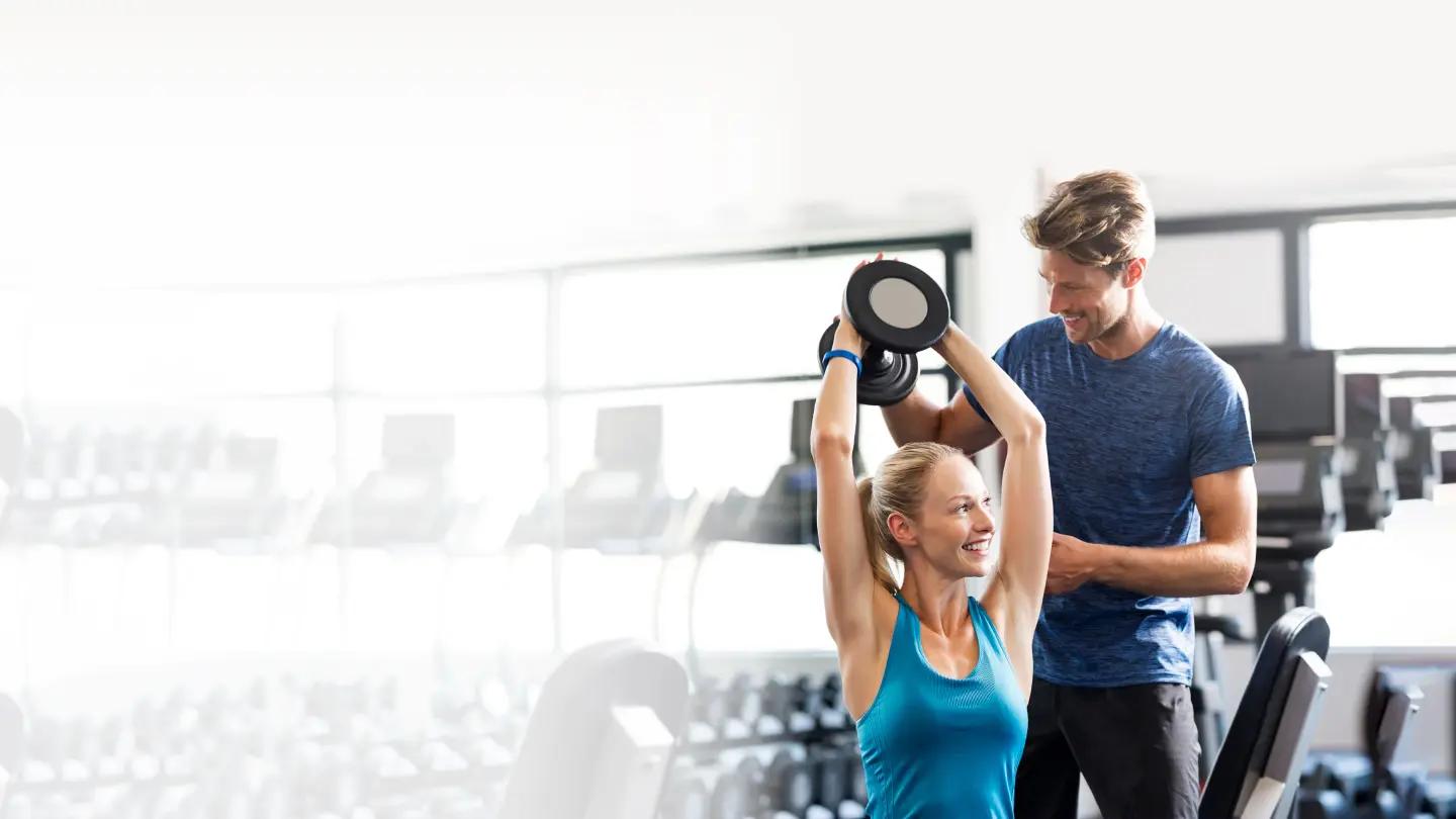 Woman being assisted by personal trainer while doing tricep presses with a dumbbell